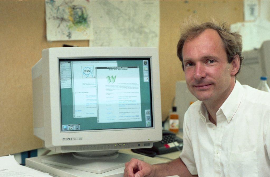 Tim Berners Lee con su ordenador a su espaldas, el cual muestra varias ventanas del sistema operativo.